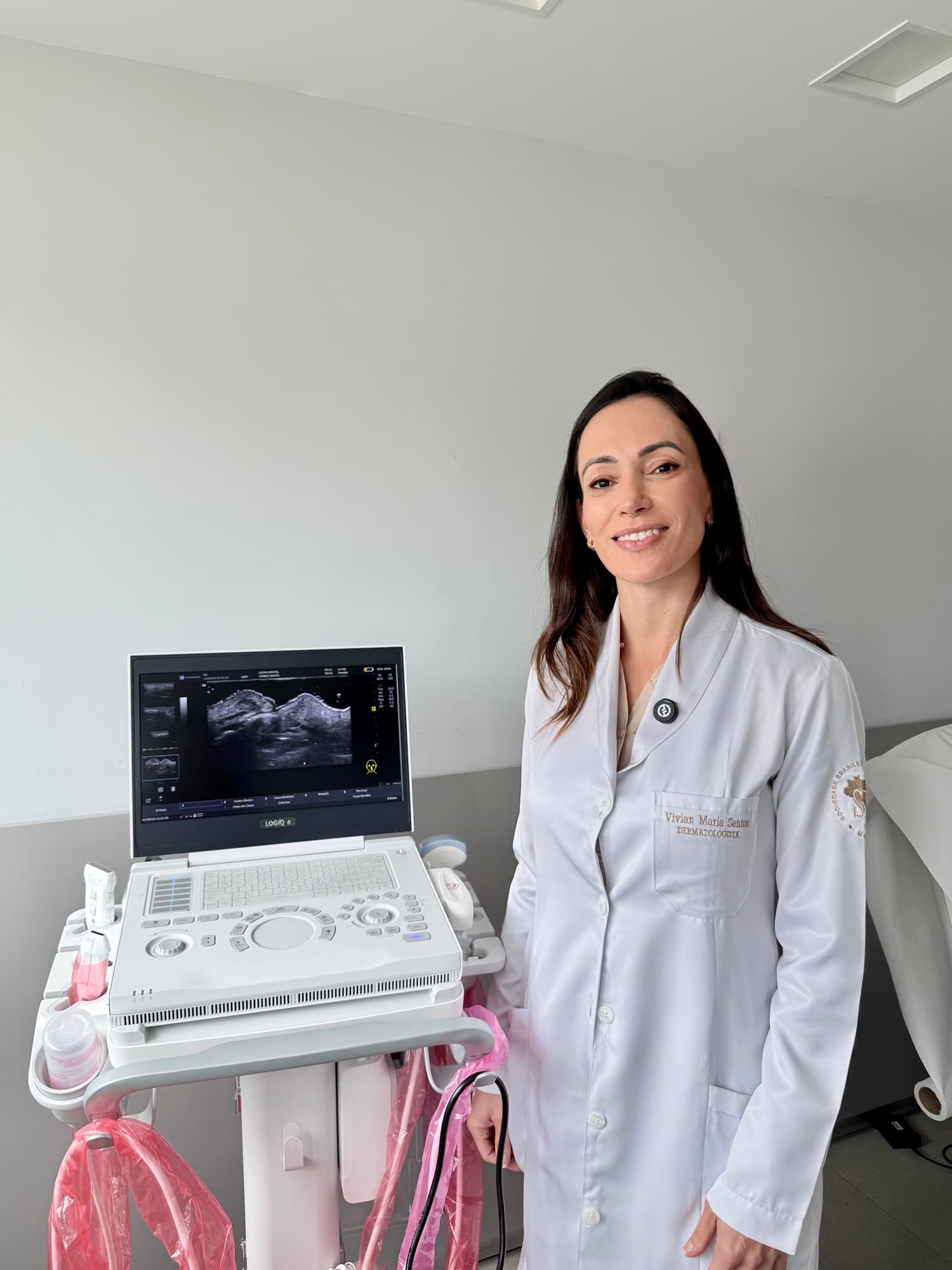 Dra. Vivian Santos, de jaleco branco, ao lado do aparelho de ultrassom dermatológico com uma imagem da pele no monitor